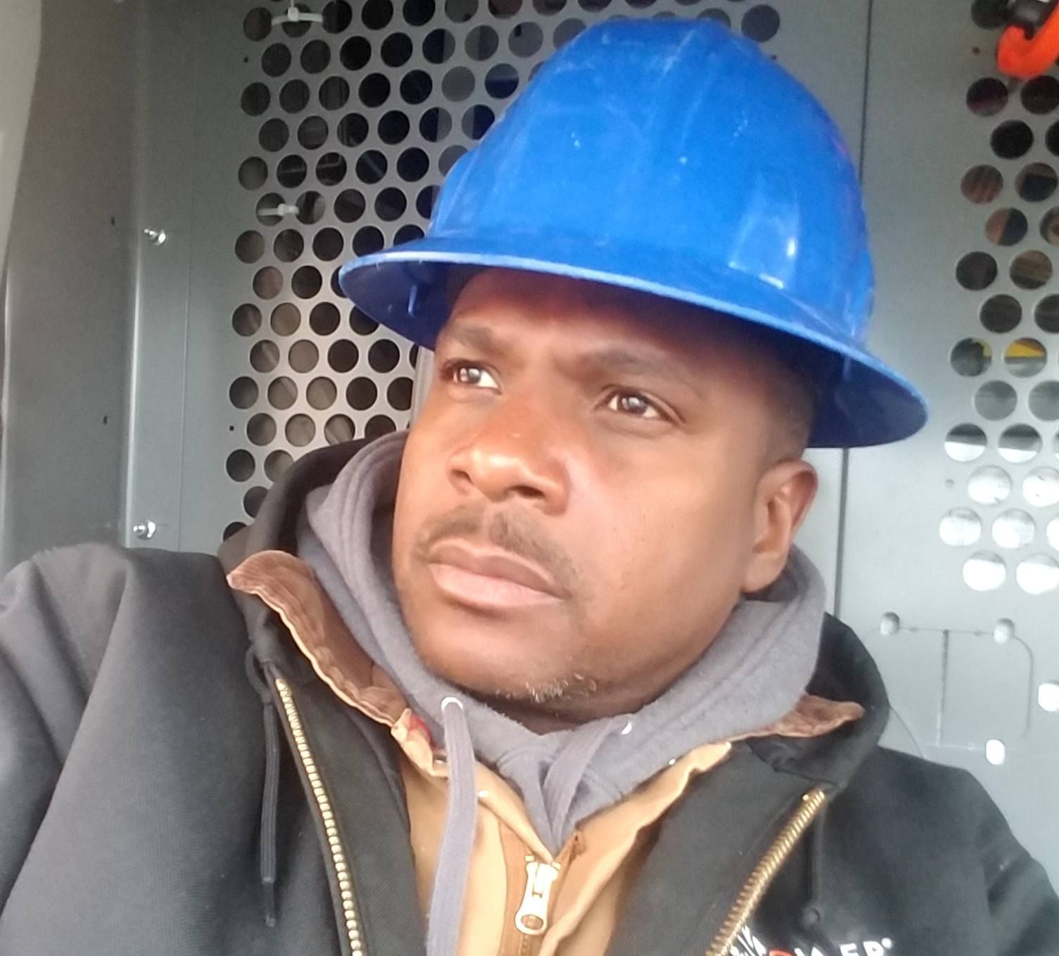 Man wearing a blue hard hat and work jacket, looking away. Inside a vehicle with metal paneling.