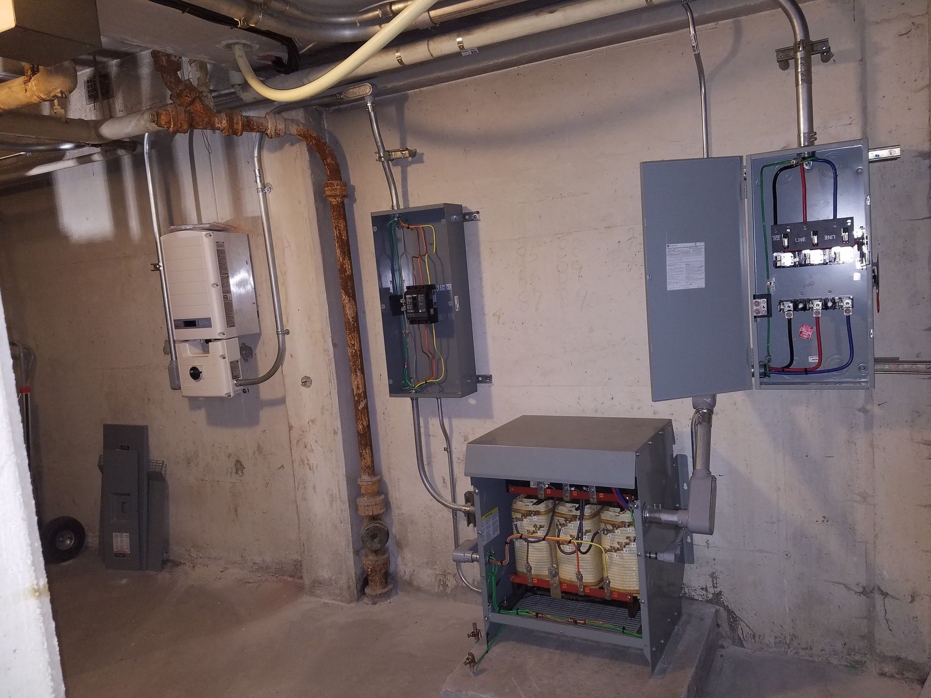 Electrical equipment in a concrete basement.  Various gray metal boxes and conduits against a light-colored wall.