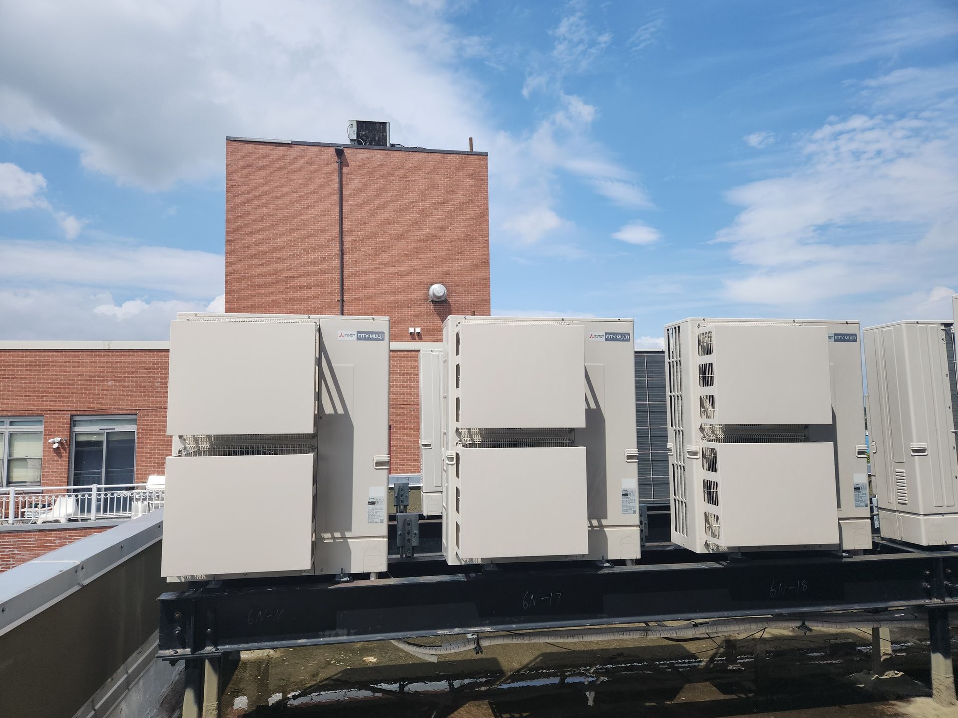 Rooftop view of several large, beige air conditioning units atop a dark surface with a brick building in the background.