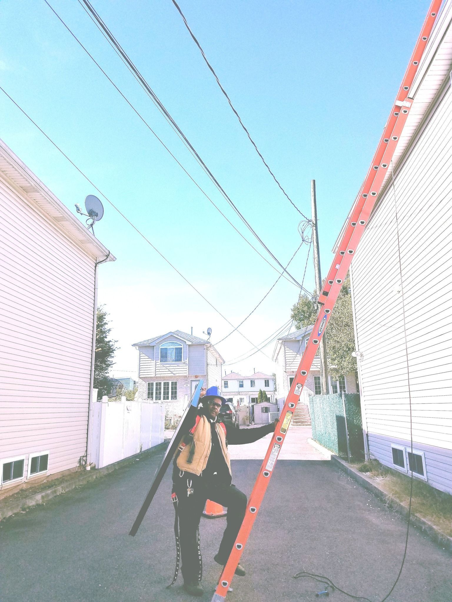 A person in safety gear steadies a ladder angled towards overhead power lines in a narrow alleyway.