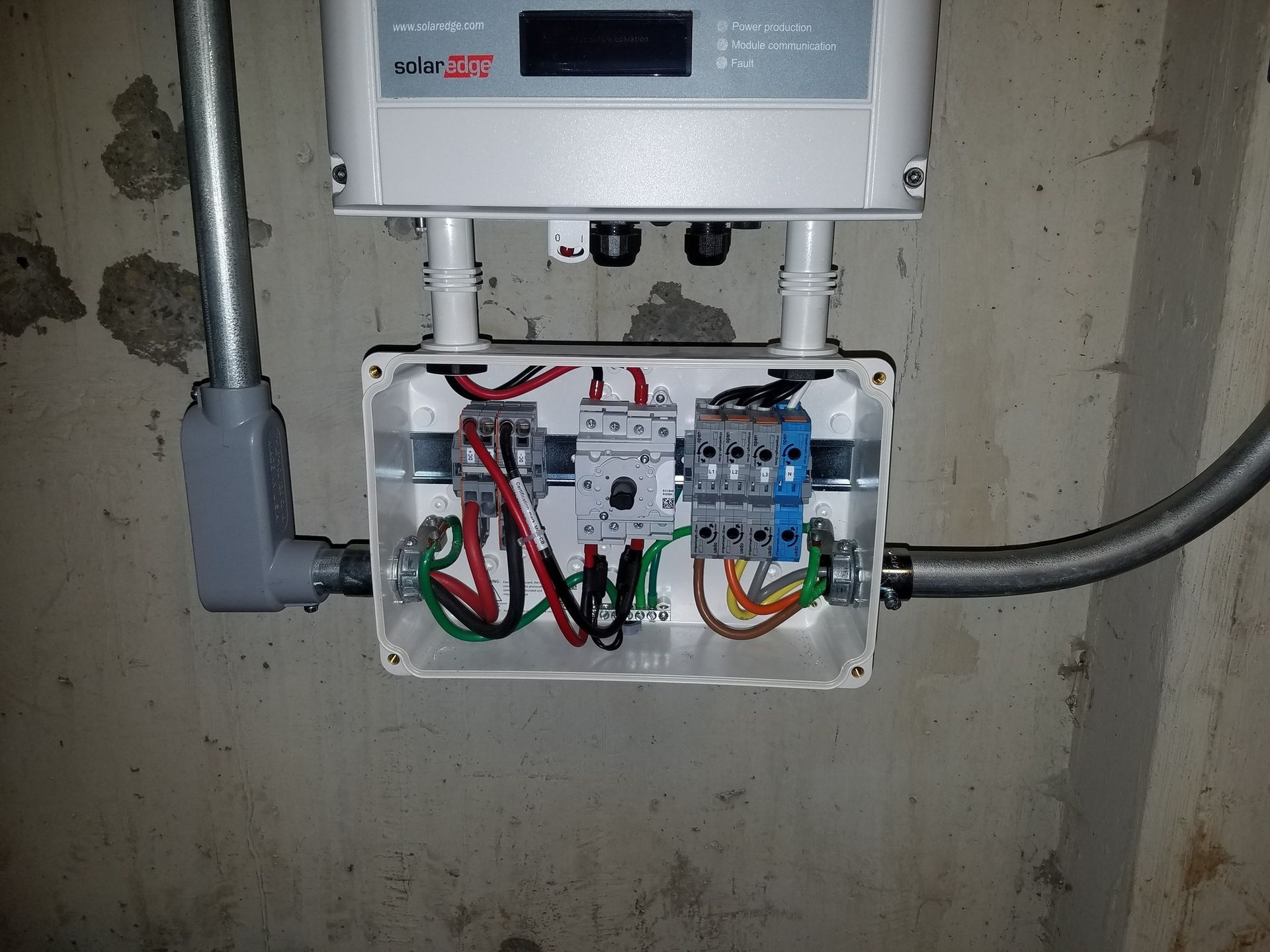 Electrical panel with wiring, conduit, and a solar inverter mounted on a concrete wall.