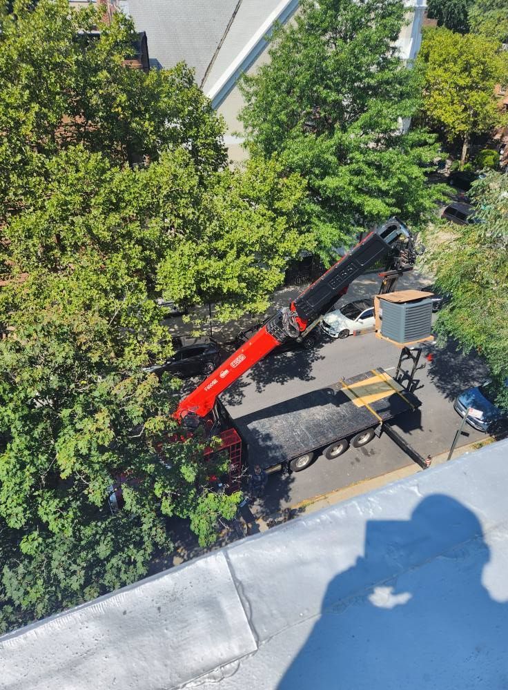 Crane extending over a flatbed truck on a street, surrounded by trees and buildings, sunny day.