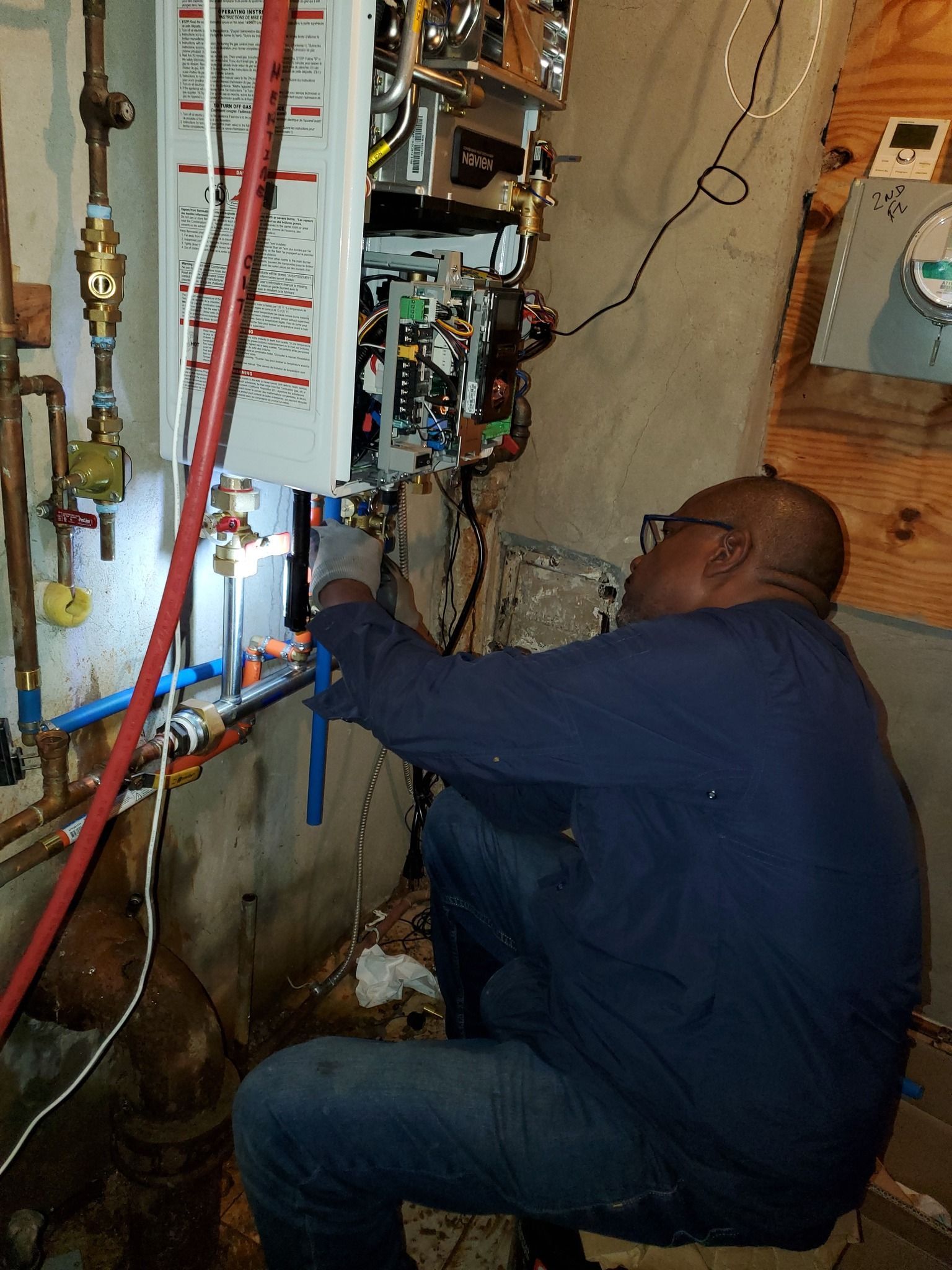 Plumber installing a water heater in a basement. Copper pipes, blue and red tubes, and wiring are visible.