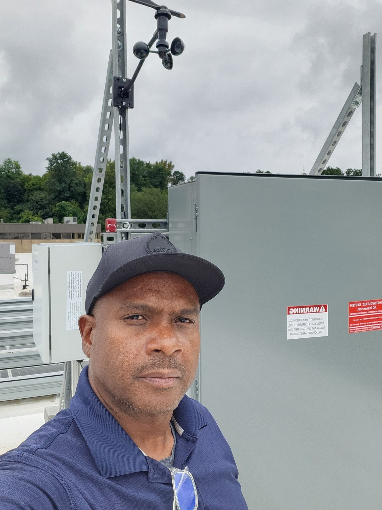 Man in a cap on a rooftop near equipment, with a weather monitoring station, under a cloudy sky.