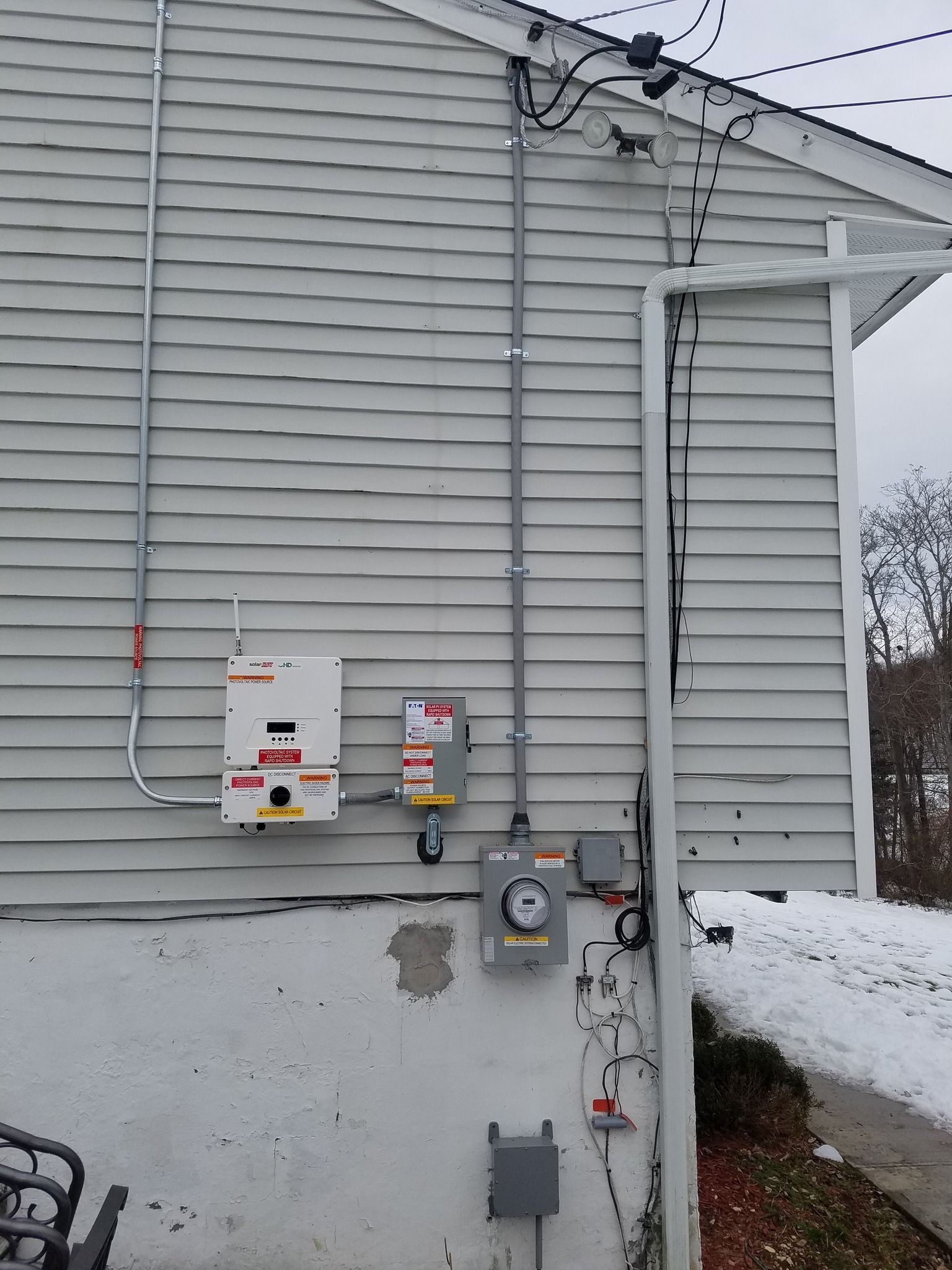 Exterior wall with electrical equipment, including a meter, panel, and conduit, on a white clapboard house.