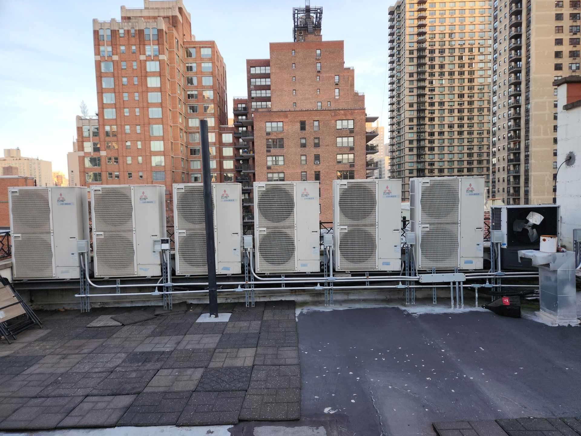 Rooftop with several air conditioning units; brick buildings in background.