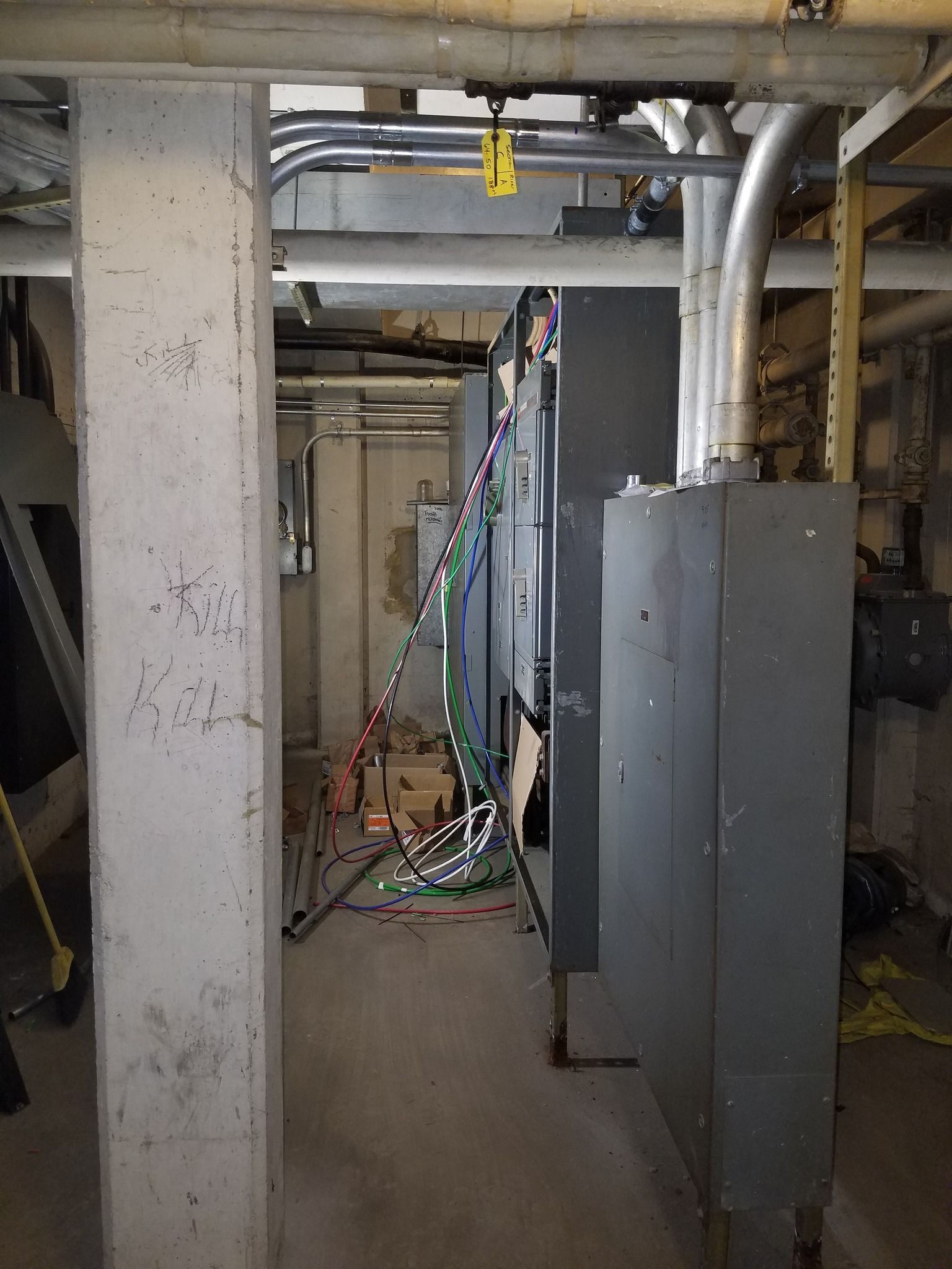 Gray electrical equipment in a narrow industrial space, with pipes above and a concrete pillar on the left.