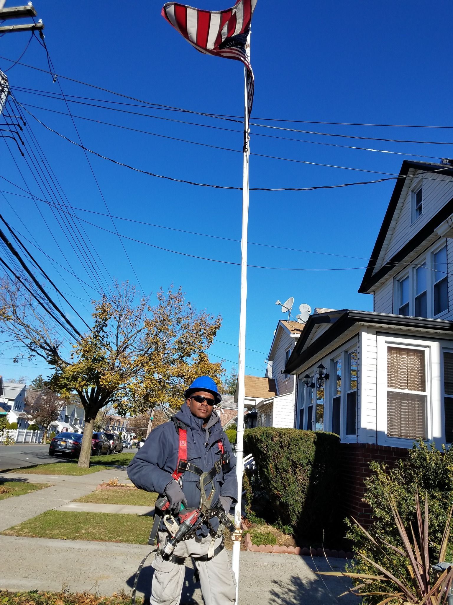 Man in work gear next to tall flag pole with a striped flag, on a sunny day in a residential area.