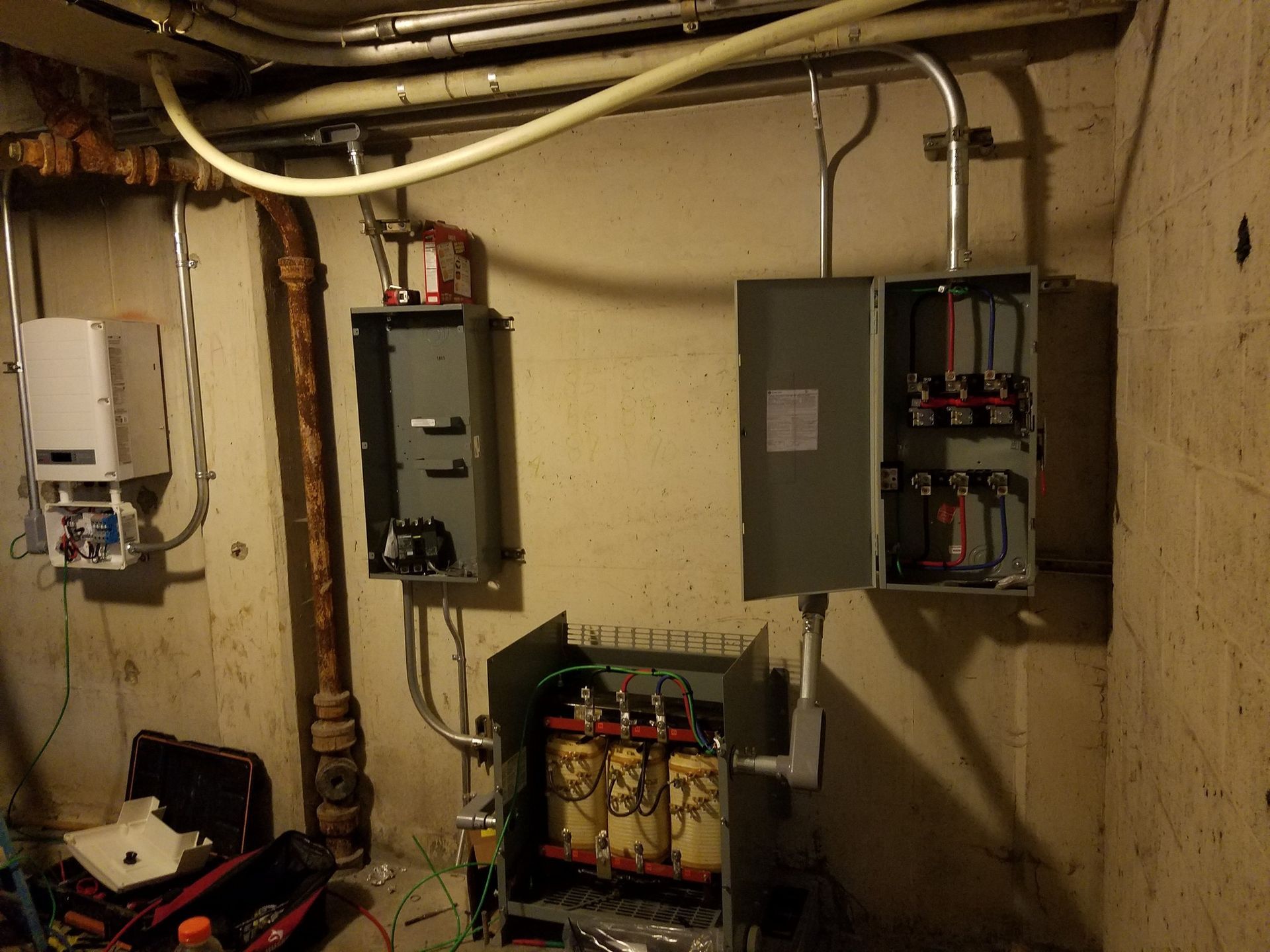 Electrical components mounted on a concrete wall. Conduit and gray metal boxes with wires, in a utility room.