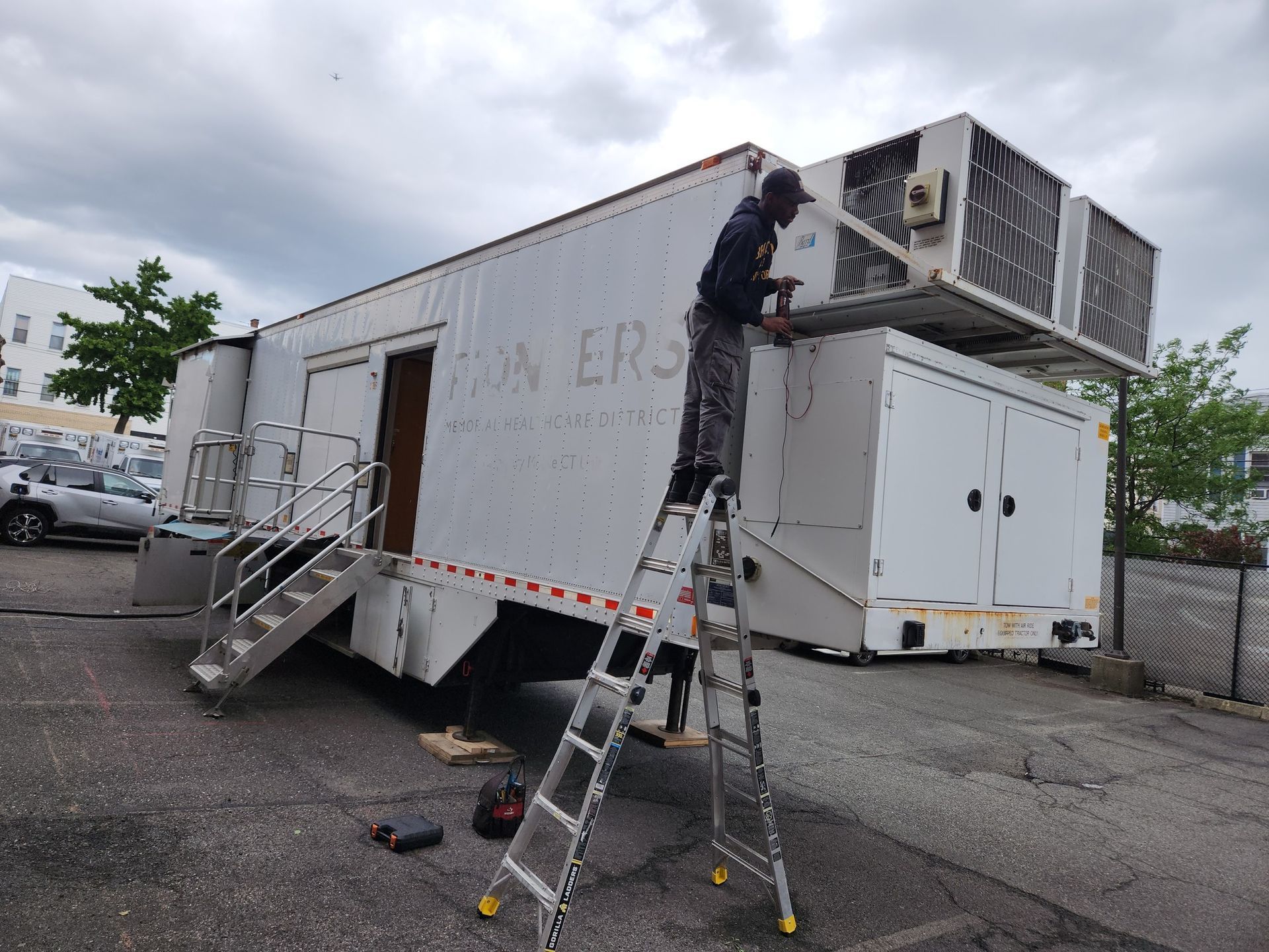 A person on a ladder working on a white trailer with equipment attached, parked outdoors.