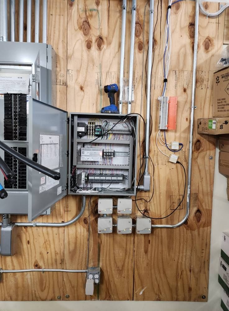 Electrical panel and conduit on plywood wall, with open control box and power outlets below.