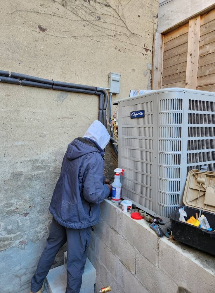 Person in a hooded jacket working on an air conditioning unit next to a building wall.