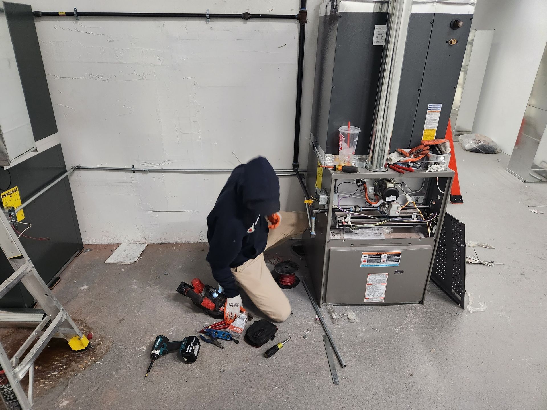 HVAC technician kneels on a concrete floor, working on an open furnace, tools scattered around.