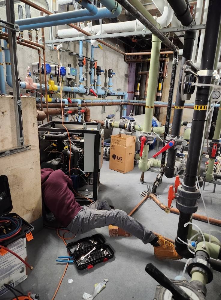 A person repairs machinery in a room with numerous pipes. The person is working on the machinery near a toolbox on the floor.