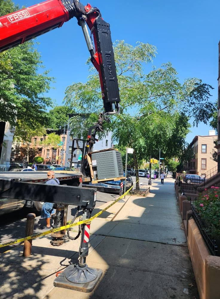 Crane lifting a large, dark object on a city street; yellow caution tape; sunny day.
