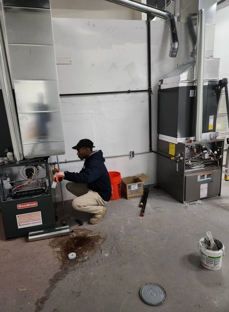 Person crouches near furnace, working on it. Gray and silver ductwork, white walls, concrete floor.