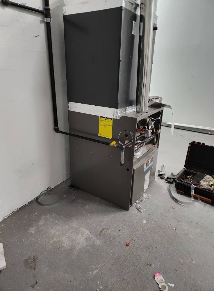 Gray furnace in a corner against white walls. Black pipe extends from the furnace. A toolbox sits on the floor.