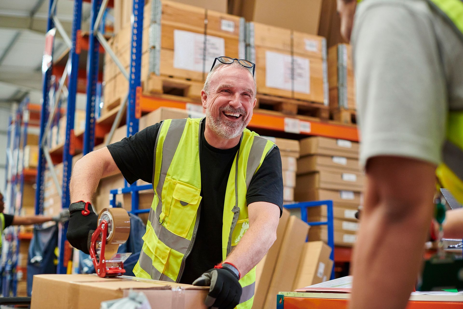Warehouse worker sealing a box with tape dispenser, surrounded by shelves of packages.