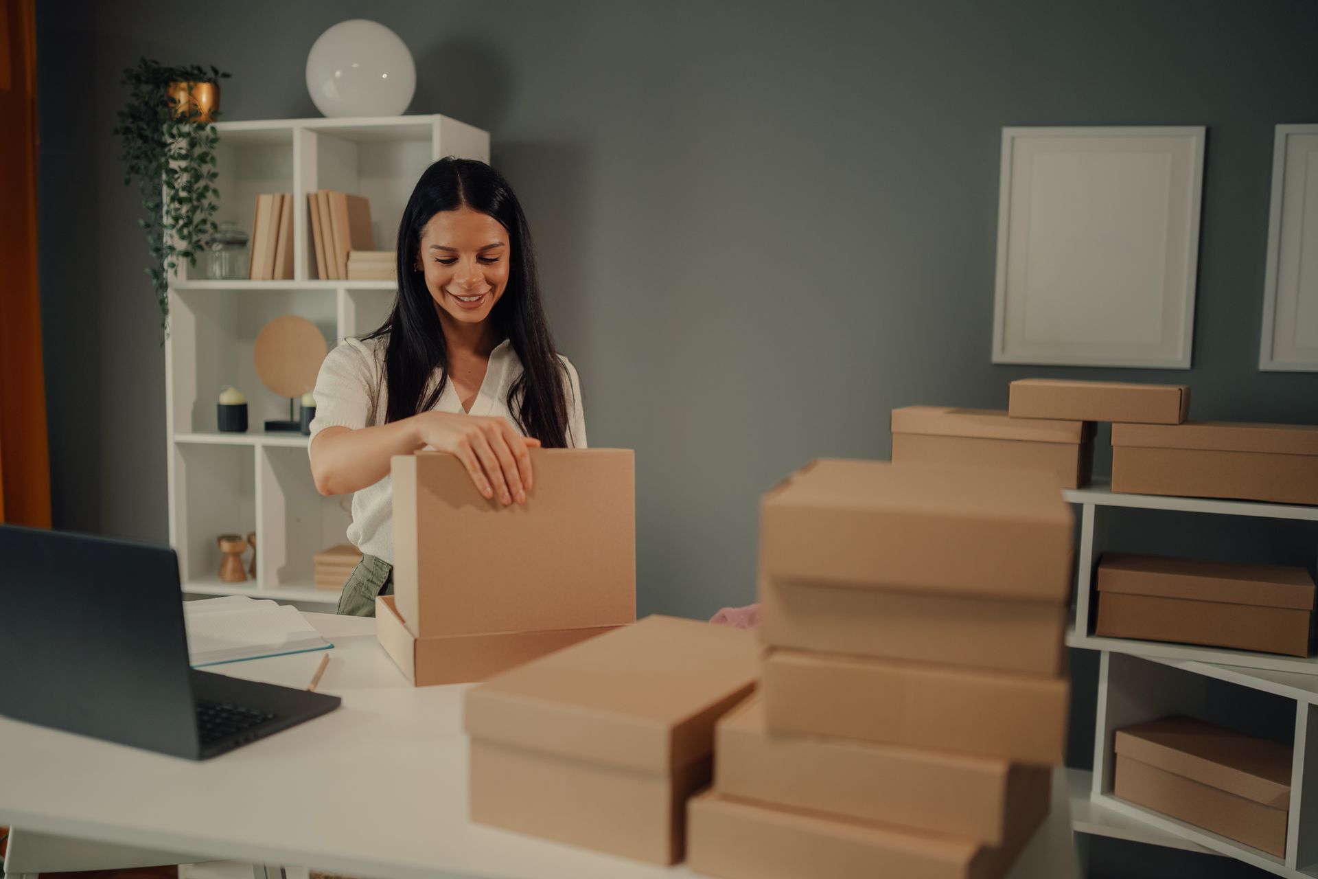 Person packing boxes in a home office with shelves and a laptop on the desk. Person packing boxes in a home office with shelves and a laptop on the desk.