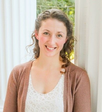 Woman smiling, wearing a brown cardigan over a white top, standing near a window.
