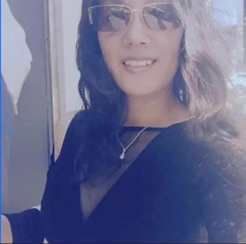 Woman wearing sunglasses, black dress, and necklace, smiling outside.