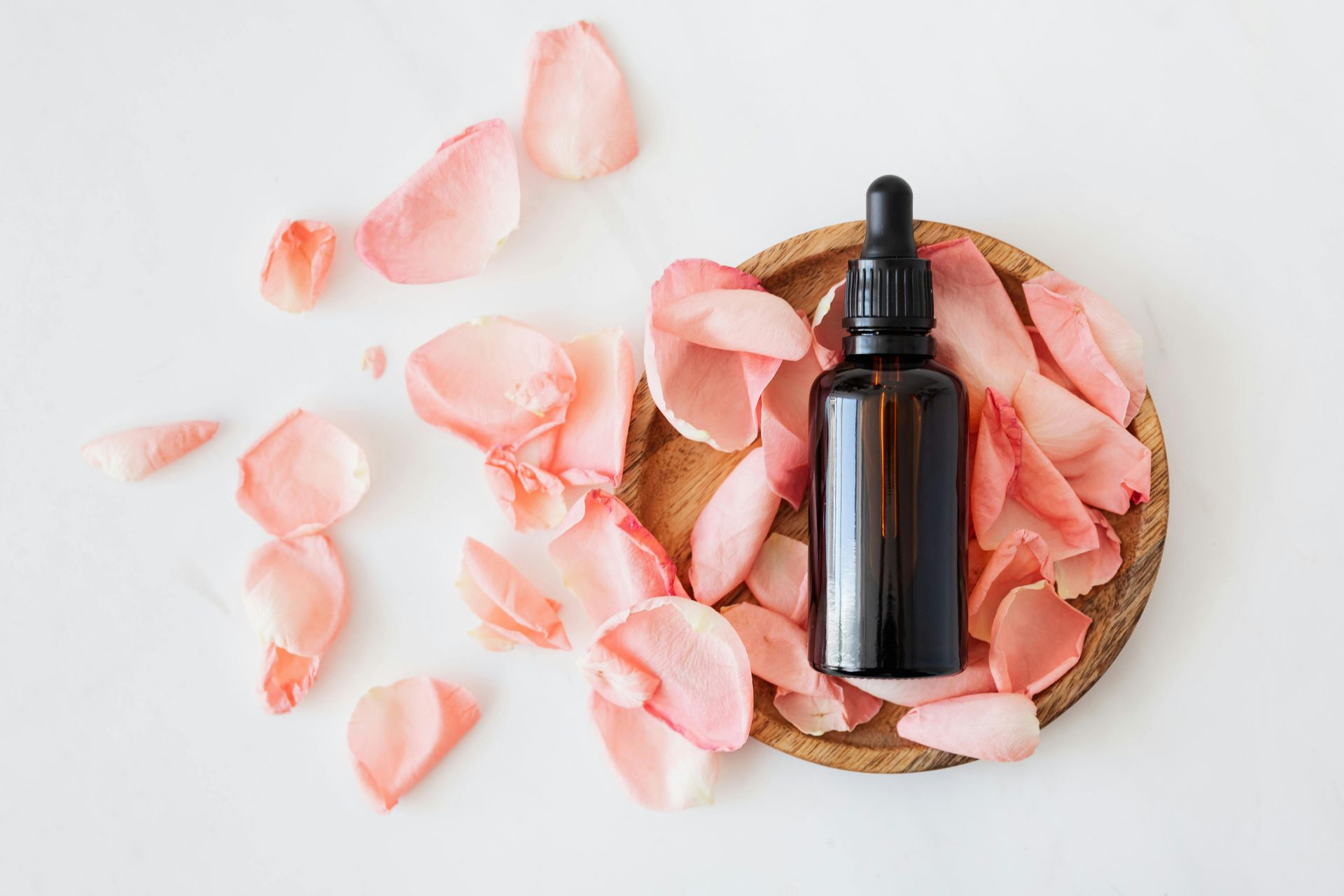 Brown bottle with dropper on wooden plate surrounded by pink rose petals, white background.
