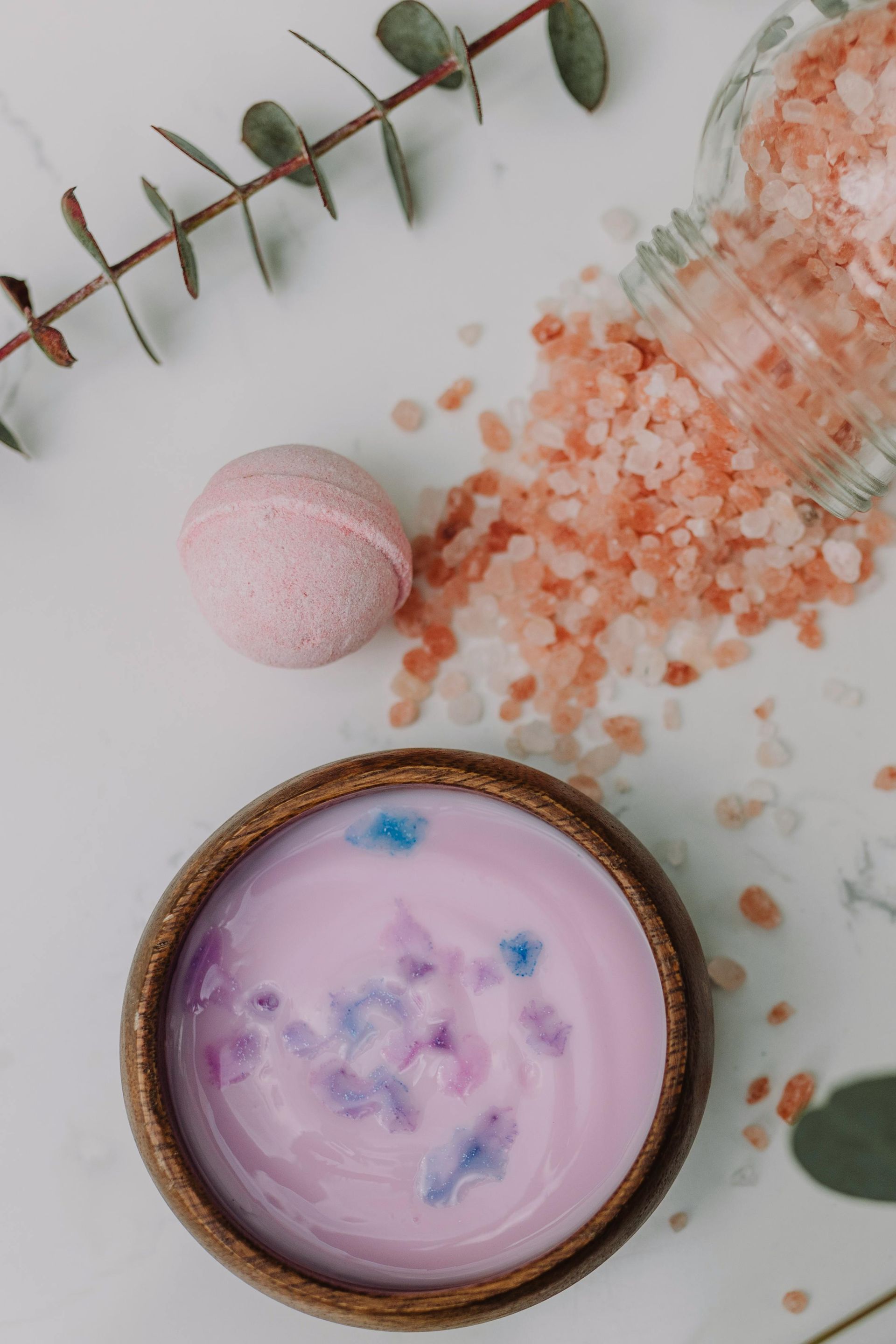Pink bath bomb and salt next to wooden bowl of pastel purple bath products, with eucalyptus.