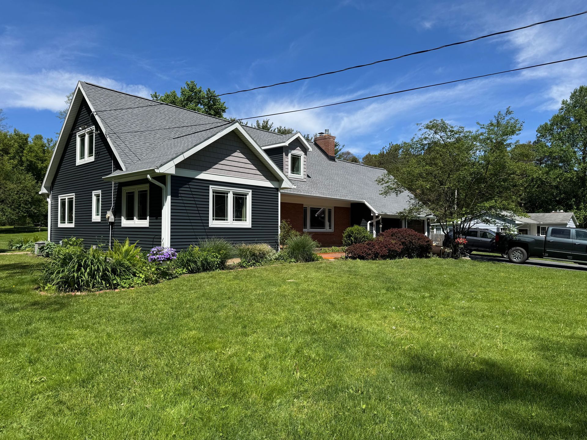 Dark house with grey roof, brick section, on green lawn. Blue sky, trees, parked vehicles in view.