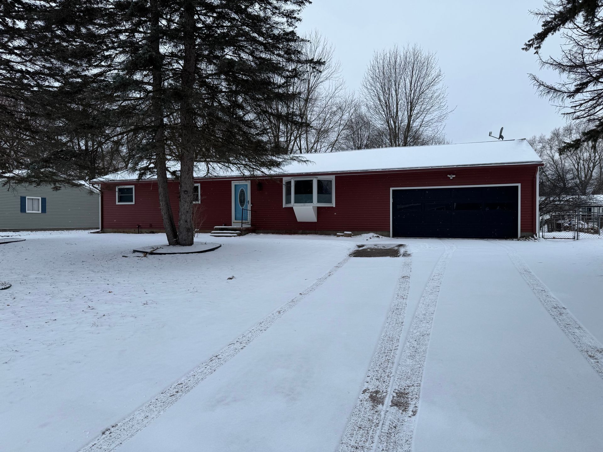 Red house with snow-covered yard and driveway; a dark garage door. Trees frame the house under a gray sky.