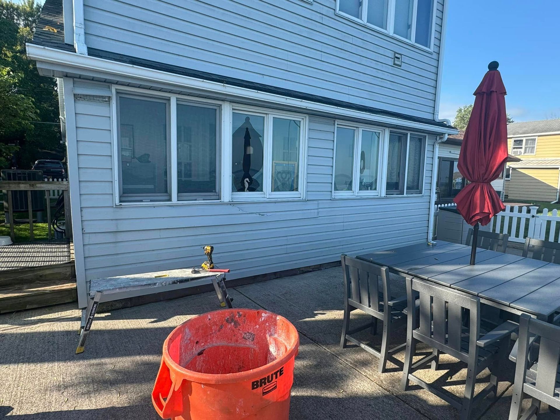 A two-story gray house with a deck, outdoor furniture, and an orange bucket. Sunny day.