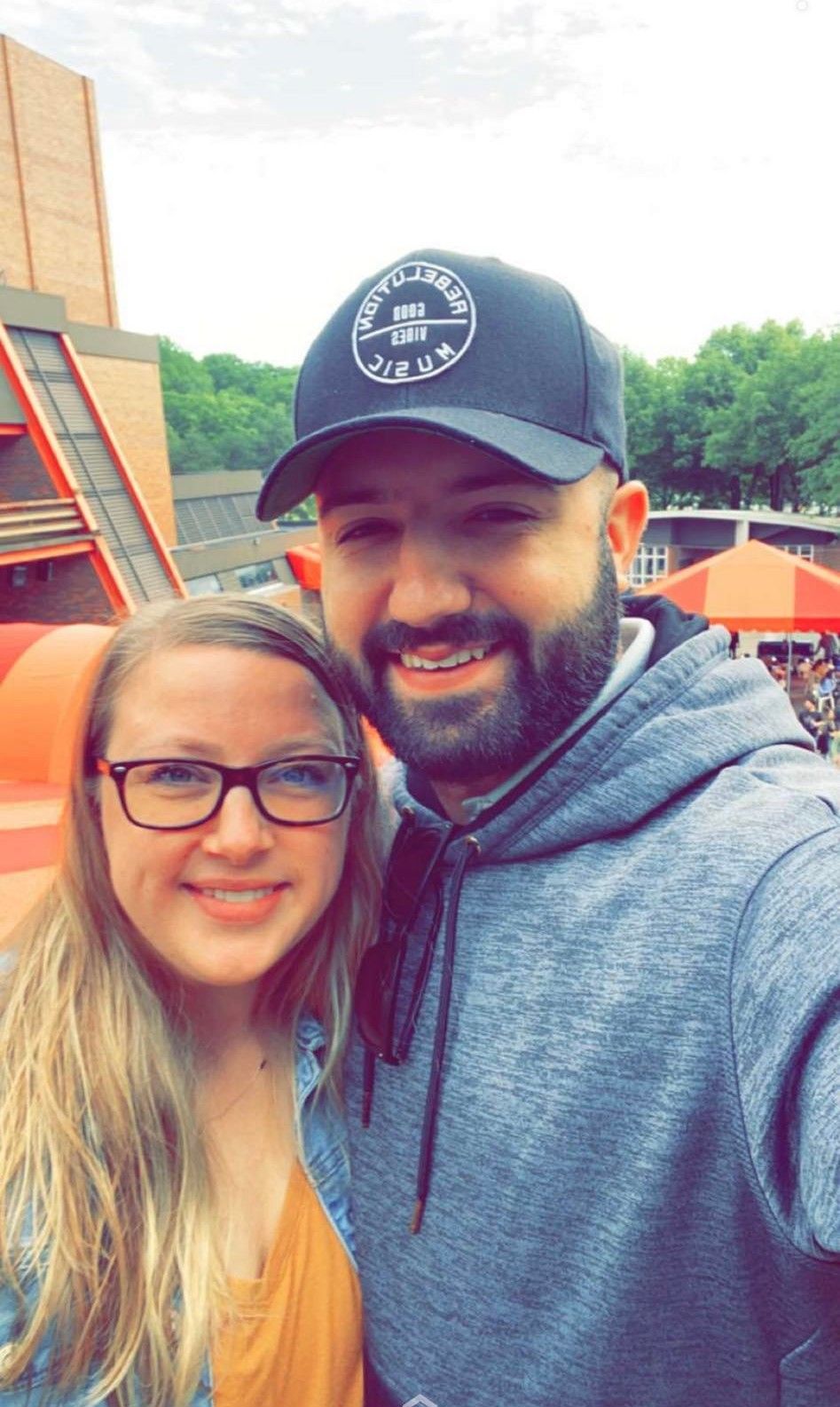 Man and woman smiling for a selfie outdoors, man wearing a hat and hoodie, woman in glasses and denim jacket.