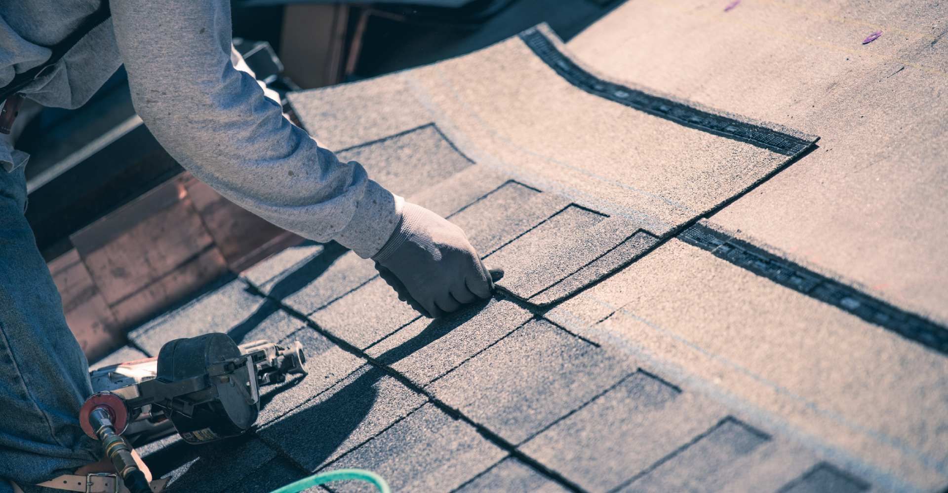 Roofer in gray shirt and gloves, installing asphalt shingles on a roof with a nail gun.