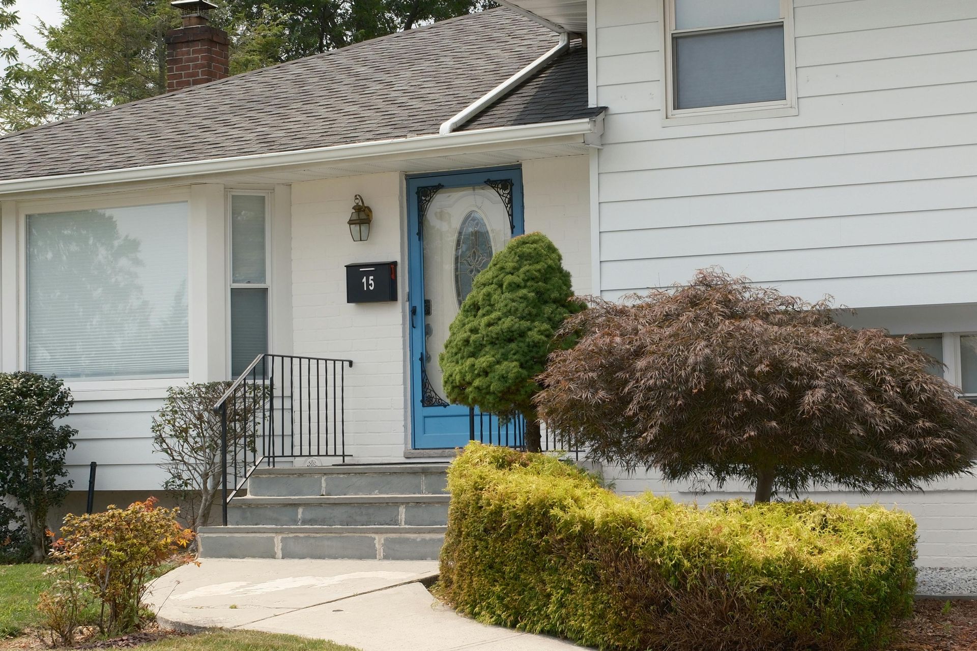 White house with blue door and steps, a brown-leafed tree and green shrub in the front yard.