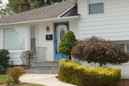 White house with blue door and steps, a brown-leafed tree and green shrub in the front yard.