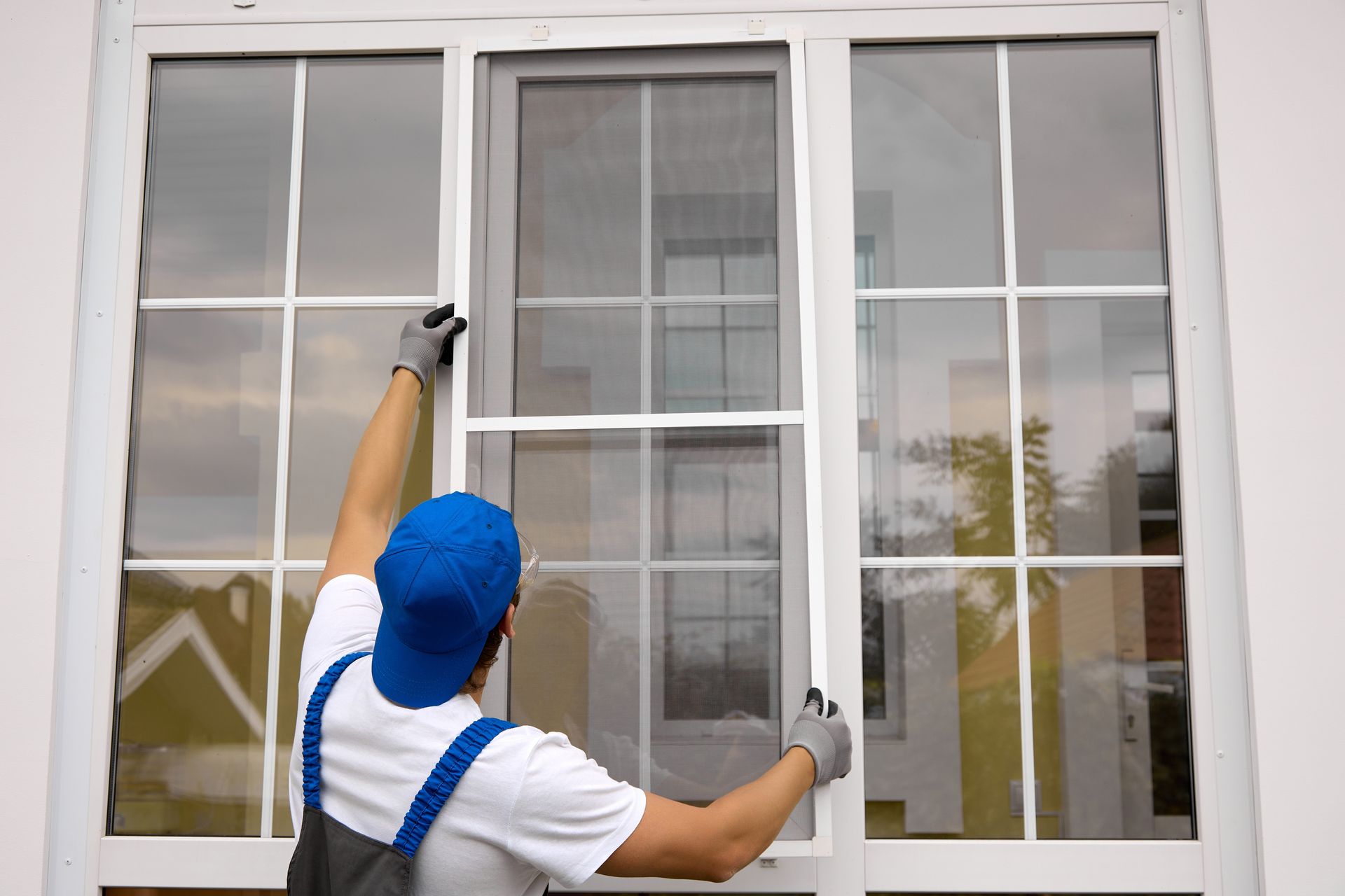 Person in blue hat installing a window screen outdoors. White window frame and building exterior.