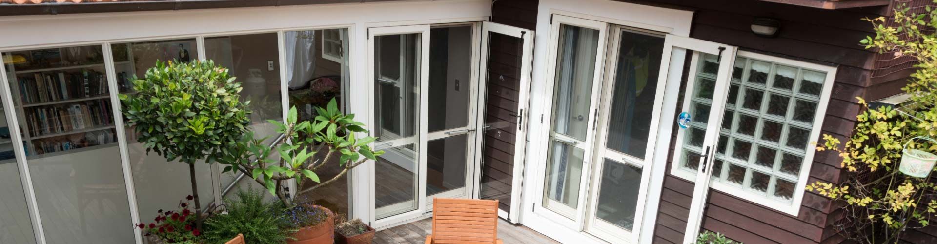 Brown house with white-framed windows, potted plants, and green foliage.