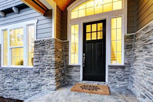 Gray and stone facade home entrance with black door, sidelights, doormat, and exterior lights.