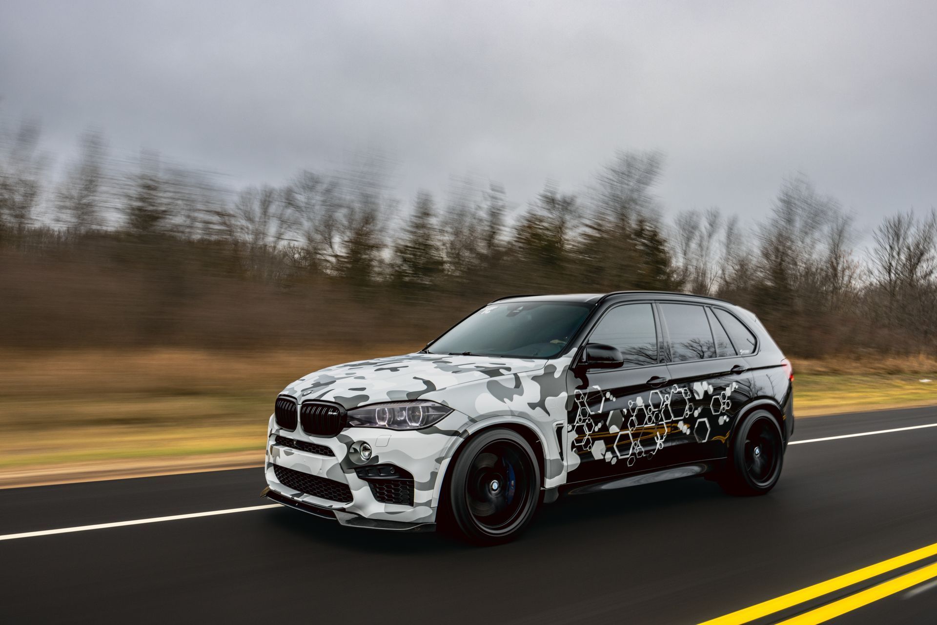 A camouflaged BMW X5 m is driving down a road.