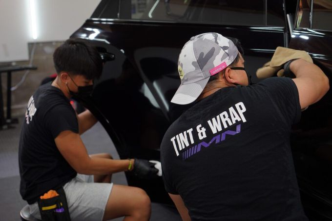 Two men are working on a car and one of them is wearing a shirt that says Tint & Wrap.