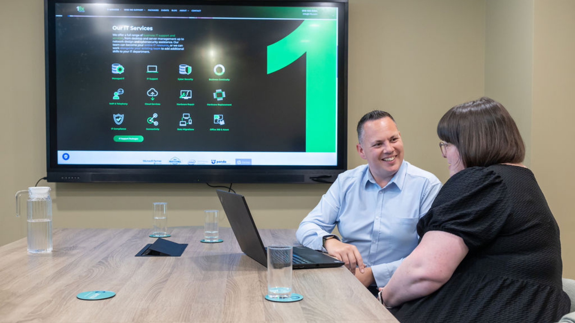 a man and a women sat smiling at each other on a desk with a laptop and screen in front of them 