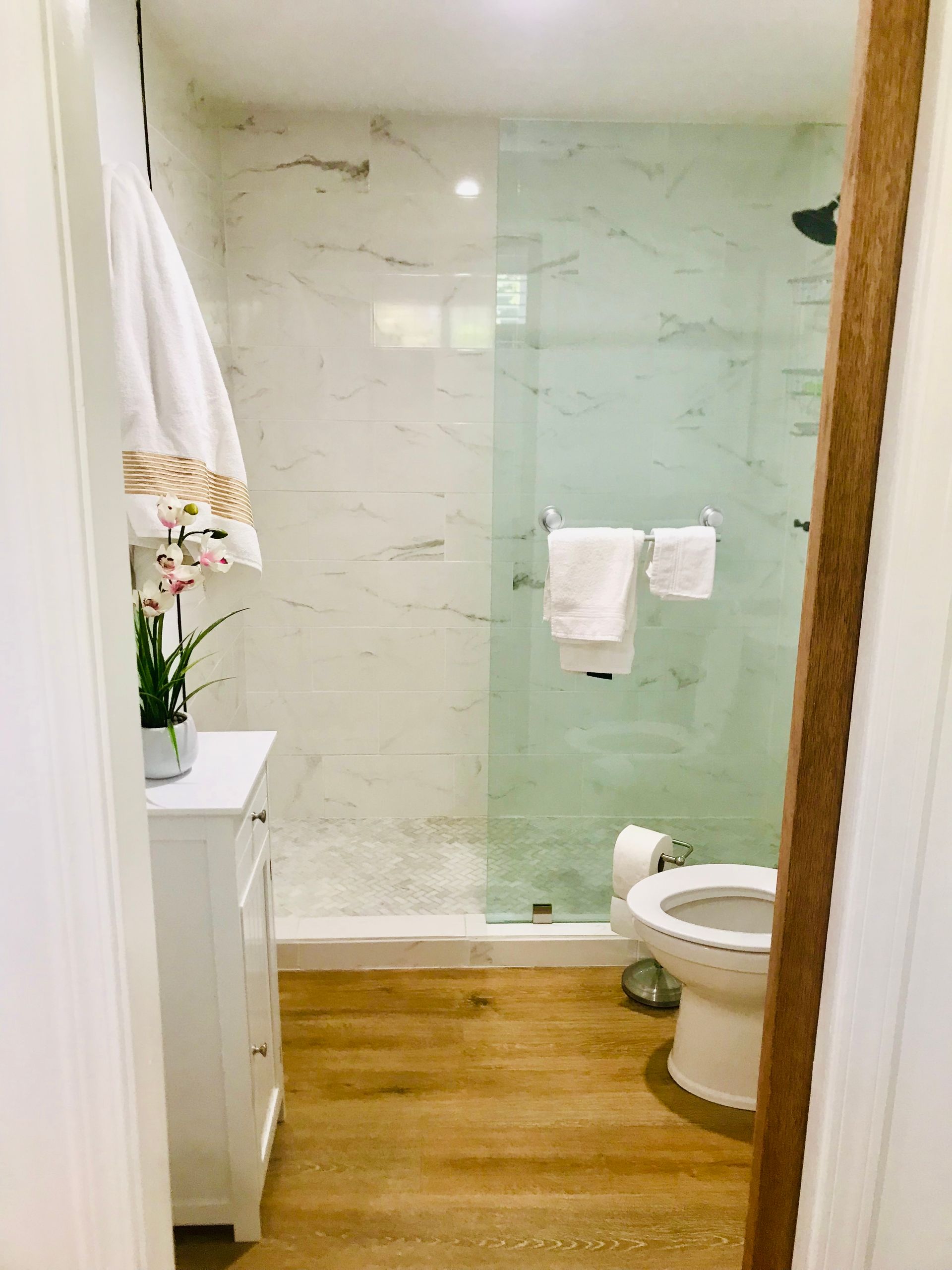 Bathroom with white marble tile, glass shower door, white vanity, and wooden floor.