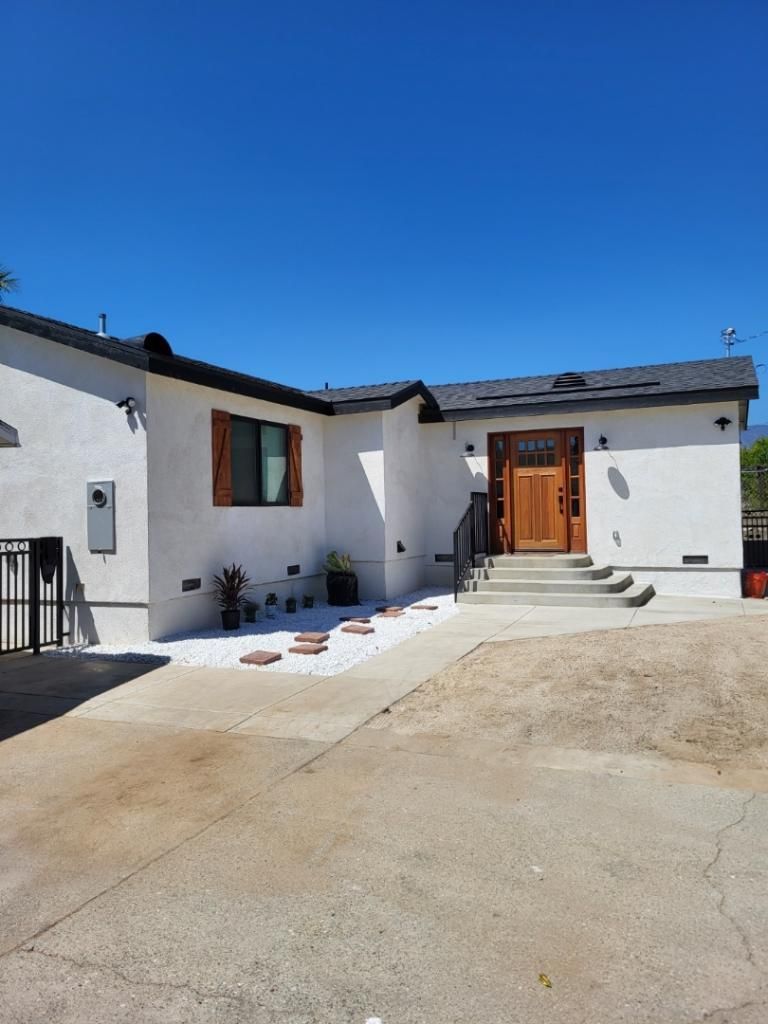 White house with brown door and shutters, concrete path, clear blue sky.