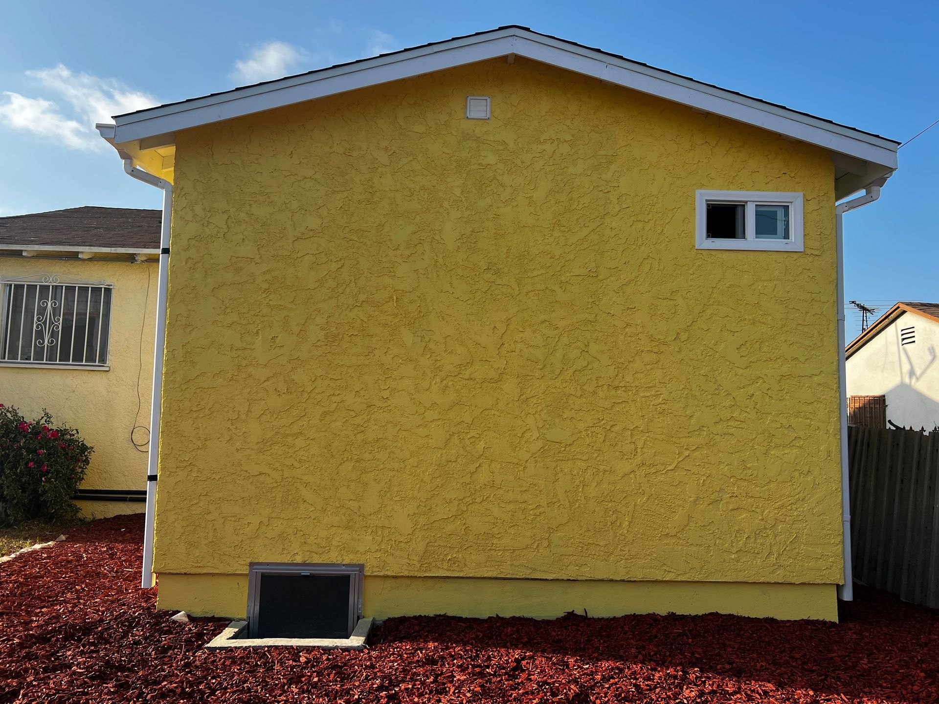 Yellow stucco house with a small window and dark window well, trimmed with white and red mulch.