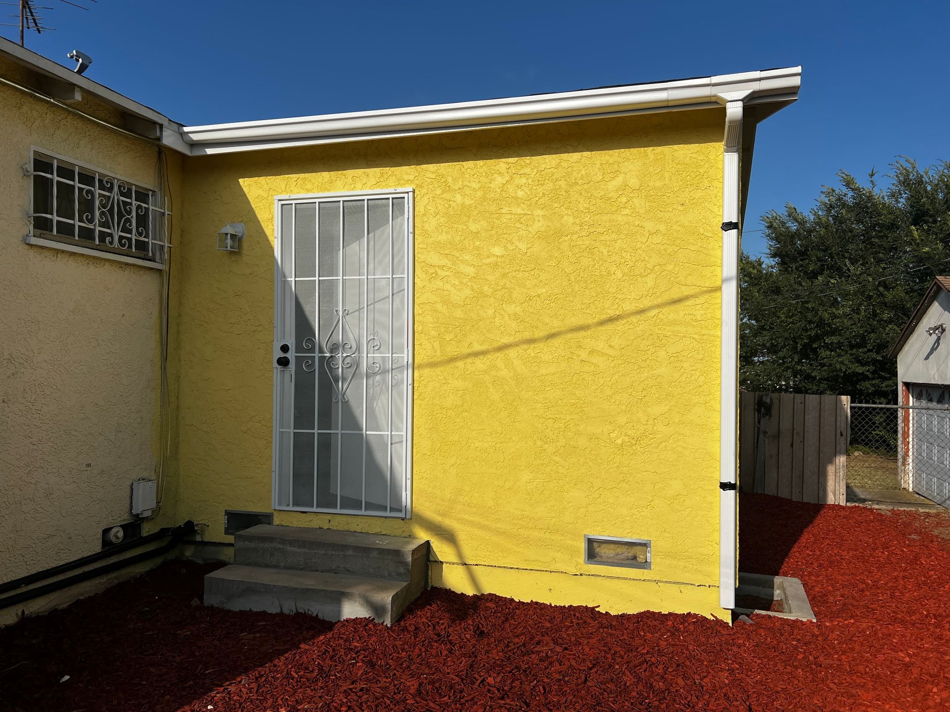 Yellow building with a security door, steps, and a gutter. Red mulch.
