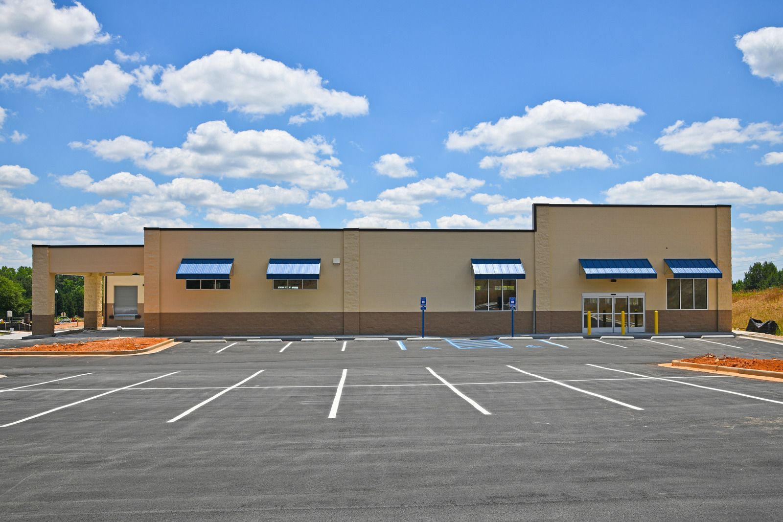 A beige, modern building with blue awnings and empty parking spaces under a blue sky.