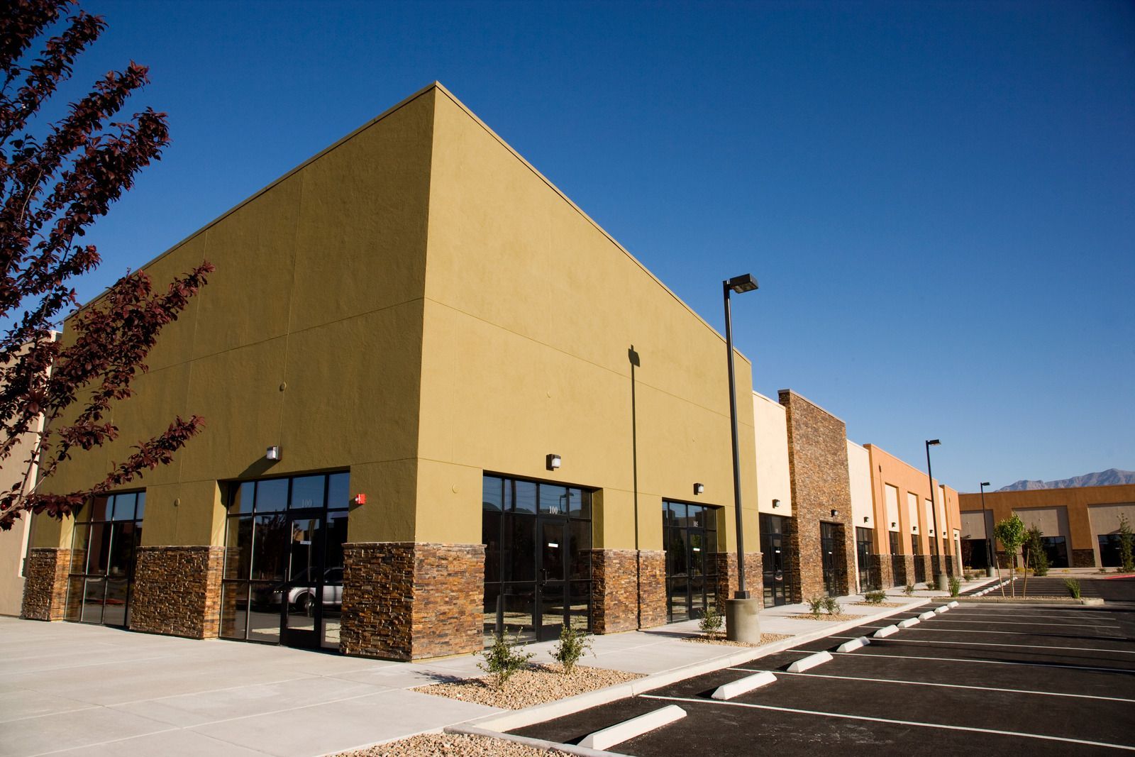 A row of retail spaces with tan and stone exteriors, glass doors, and a parking lot under a blue sky.