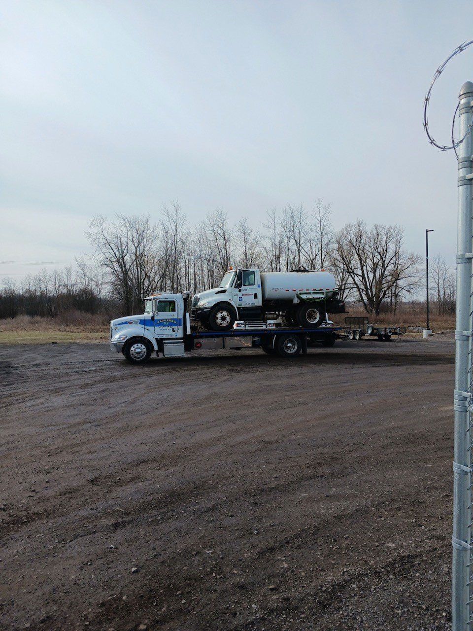 Truck hauling a water tanker truck on a gravel lot under a cloudy sky.
