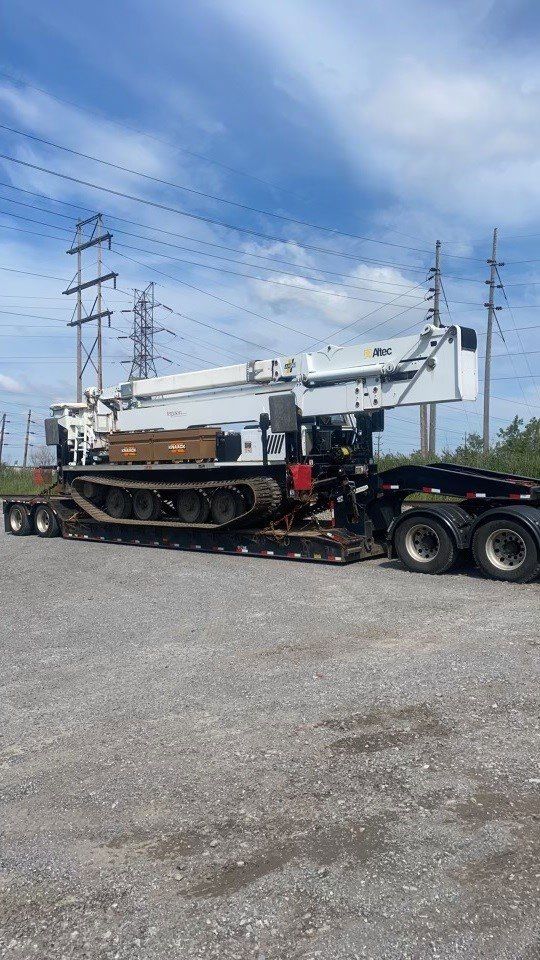 Heavy white tracked crane on a flatbed trailer, under power lines, gravel lot, blue sky.