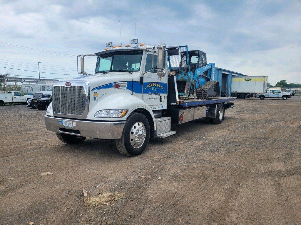 White and blue tow truck with a blue construction vehicle on the flatbed in a gravel lot.