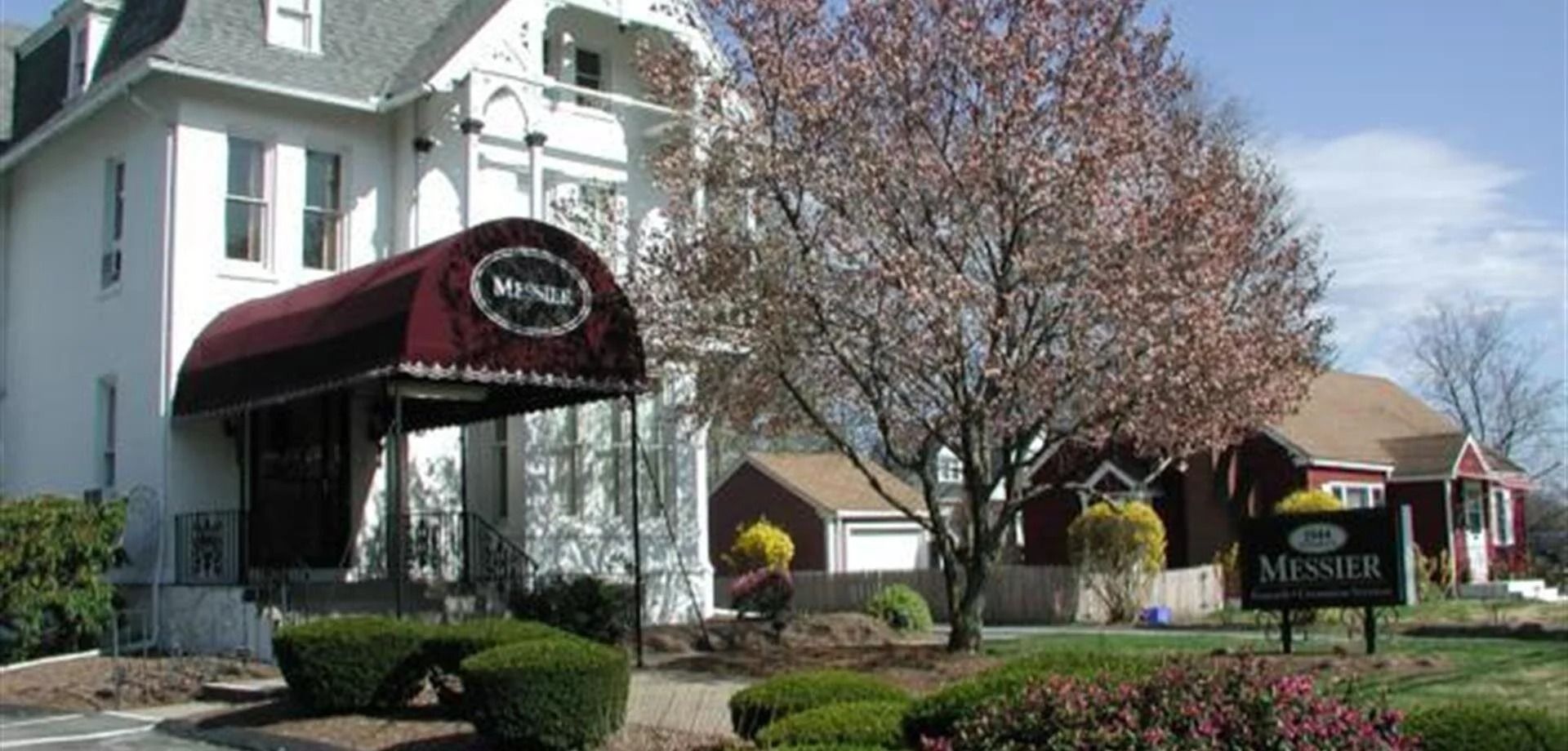 Exterior view of a white building with a maroon awning, next to a tree with pink blossoms and other buildings with a sign that says 